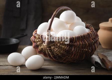 Korbkorb mit weißen Hühnereiern auf Holztisch. Bratpfanne und Tontopf auf dem Hintergrund. Stockfoto