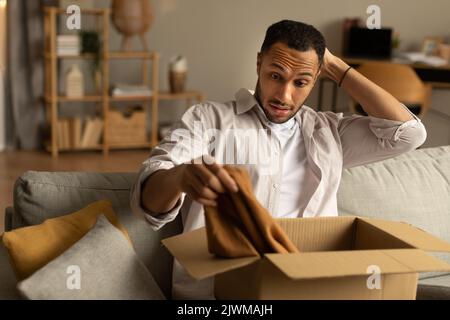 Unzufriedener Käufer. Frustrierter schwarzer Mann, der das Paket nach der Lieferung auspackend auspackt, Kleidung hält und zu Hause auf der Couch sitzt Stockfoto