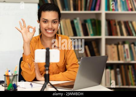 E-Learning. Positive Lehrerin, die Online-Unterricht gibt, eine gute Geste zeigt, mit einer Smartphone-Webcam spricht und lächelt Stockfoto