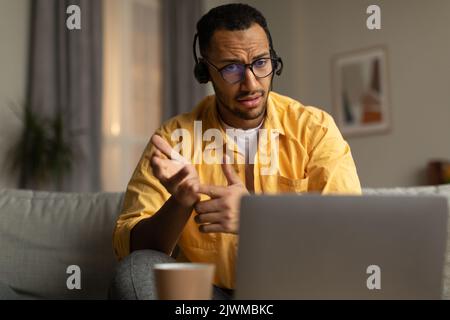 Seriöser junger schwarzer Mann mit Kopfhörern, der im Heimbüro eine Webkonferenz oder ein entferntes Geschäftstreffen auf einem Laptop abhalten kann Stockfoto