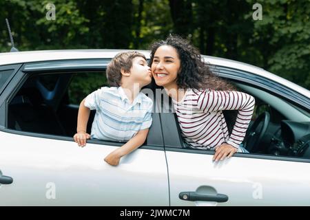 Fröhlicher kleiner arabischer Junge küsst seine Mutter auf die Wange durch offenes Fenster im neuen Auto, genießen Sie die gemeinsame Reise Stockfoto