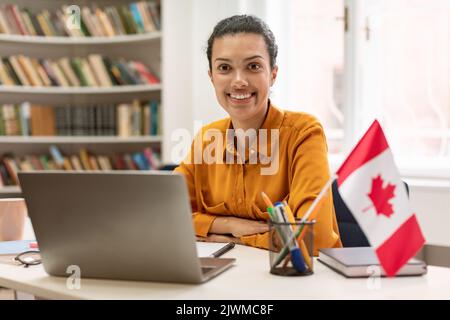 Online Fremdsprachen Nachhilfe. Glückliche Lehrerin, die mit der Flagge Kanadas in der Bibliothek sitzt und einen Laptop benutzt Stockfoto