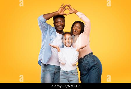 Aufgeregte schwarze Eltern zeigen Herzform mit den Fingern und bilden ein symbolisches Dach unter dem Kopf ihrer Tochter Stockfoto