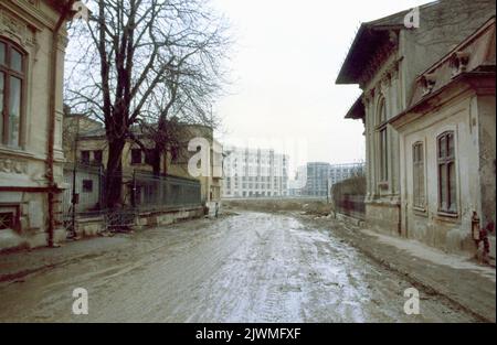 Bukarest, Rumänien, Januar 1990, wenige Wochen nach dem Fall des Kommunismus. Als Ceausescu seinen Schwerpunkt auf dem Bau seiner Megalomanischen Projekte wie dem Civic Center legte, verschlechterten sich die historischen Viertel der Hauptstadt langsam. Einige historische Häuser in der Nähe des Victory of Socialism Blvd. Wurden für den Abriss evakuiert und entkamen ihm dank der antikommunistischen Revolution kaum. Auf diesem Bild eine schlammige Straße im alten Stadtteil '11 Junie' mit Blick auf die neuen imposanten Gebäude entlang des Boulevard des Sieges des Sozialismus. Stockfoto