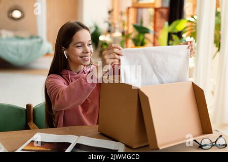 Zufriedener Kunde. Happy teen Mädchen öffnen und Auspacken Paket Karton-Box, sitzen am Tisch zu Hause und lächelnd Stockfoto