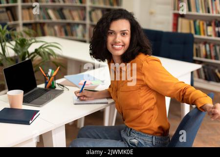 Arbeiten an einem Forschungsprojekt. Glückliche Schülerin, die in der Bibliothek einen Laptop benutzt, in einem Notizblock schreibt und die Kamera anlächelt Stockfoto