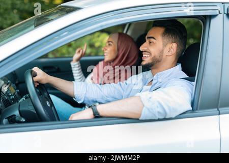 Fröhlich lächelnder, tausendjähriger islamischer Ehemann, der mit seiner Frau im Hijab am Steuer fährt, im Auto fährt, Musik genießt Stockfoto