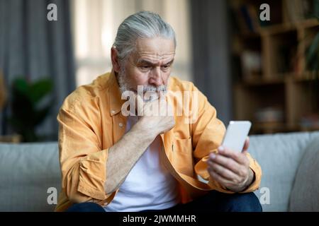 Nachdenklicher älterer Mann, der mit dem Smartphone auf der Couch sitzt Stockfoto