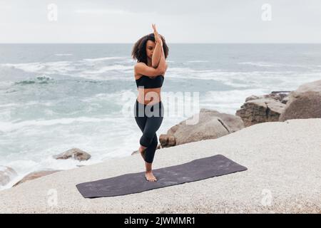 Frau, die auf einer Matte in Garudasana-Pose am Meer steht. Junge Hündin mit lockigen Haaren übt Adlerpose. Stockfoto