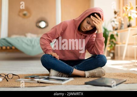 Exhautsed Teenager Mädchen macht Hausaufgaben allein, sitzen auf dem Boden mit Laptop und Buch, berühren ihren Kopf Stockfoto