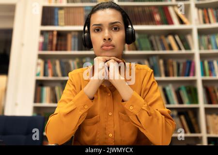 Porträtansicht einer ernsthaften Studentin in Kopfhörern, die einen Laptop anschaut und im Inneren der Bibliothek online studiert Stockfoto