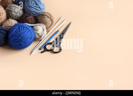 Blaue, graue und braune Strickgarnkugeln mit Schere, Häkel- und Stricknadeln auf beigem Hintergrund für Hobbys. Stockfoto