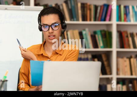 Online-Schulungen. Junge Lehrerin mit Videokonferenz mit Studenten, im Gespräch mit Laptop-Webcam, in der Bibliothek sitzend Stockfoto