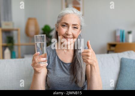 Glückliche hübsche kaukasische ältere grauhaarige Hündin hält ein Glas Wasser und zeigt den Daumen nach oben Stockfoto