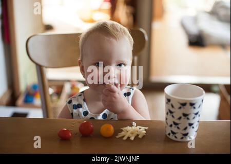 Ein 18 Monate altes Baby sitzt am Tisch und genießt einen Snack mit Kirschtomaten, geriebenem Käse und einem Getränk Wasser. Stockfoto