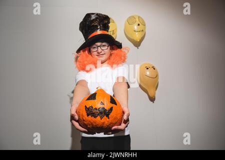 Defokussieren Sie den Vordergrund. Frohe Halloween. Kind in oranger Perücke und schwarzem Hut hält Kürbis mit gemaltem Gesicht. Das Mädchen lächelt und ist müde Stockfoto