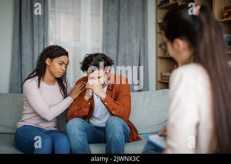 Traurige, tausendjährige schwarze Dame beruhigt den europäischen Mann bei der Begegnung mit dem Psychologen im Inneren der Büroklinik, im Kopierraum Stockfoto