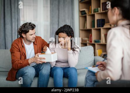 Unglücklicher kaukasischer Ehemann beruhigt und unterstützt weinende schwarze Frau auf Rücksprache mit dem Psychologen Stockfoto