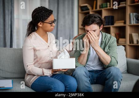 Ernsthafte, tausendjährige schwarze Psychologin beruhigt traurig depressiven europäischen Mann, Patient, der an psychischen Problemen leidet Stockfoto