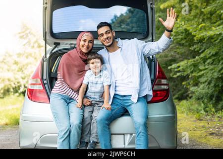 Zufriedenes tausendjähriges islamisches Männchen und Weibchen im Hijab mit Sohn, im Kofferraum sitzend und mit der Hand winkend Stockfoto