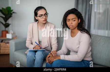 Psychiatrische Unterstützung. Traurige junge europäische Ärztin mit Brille beruhigt die schwarze Patientin im Büro Stockfoto