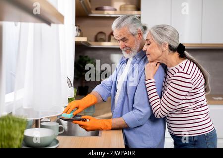 Liebevolle Ältere Frau Umarmt Ihren Mann, Während Er In Der Küche Geschirr Wäscht Stockfoto