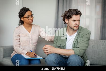 Traurige junge schwarze Ärztin beruhigt unglücklich verzweifelte kaukasische männliche Patientin im Inneren der Büroklinik Stockfoto