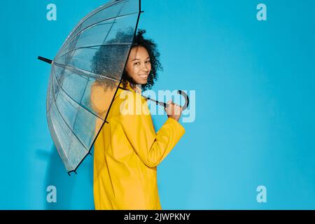 Afroamerikanische Frau in gelber wasserdichter Regenjacke mit transparentem Regenschirm isoliert auf blauem Studiohintergrund. Konzept der Herbstsaison Stockfoto