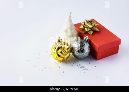 Weihnachtskomposition aus einer roten Geschenkbox mit goldener Schleife, goldenen und silbernen Weihnachtskugeln und einer weißen Weihnachtsbaumkerze auf grauem Hintergrund mit Stockfoto