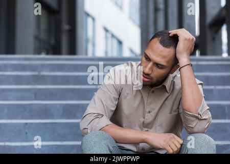 Enttäuschter und trauriger Mann, der auf einer Treppe vor dem Bürogebäude saß, ein Mitarbeiter wurde von der Arbeit entlassen, ein afroamerikanischer Mann im Hemd verärgert und depressiv. Stockfoto