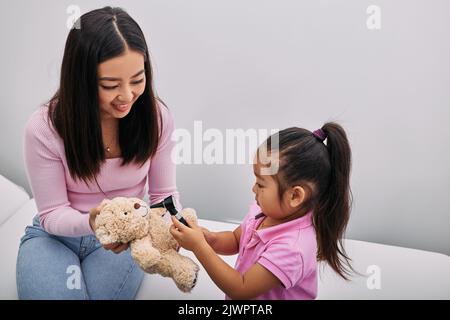 Asiatische Mädchen spielt mit ihrem Spielzeug Bär in medizinischen Spiel, mit Otoskop mit Unterstützung ihrer Mutter. Hörklinik für Kinder Stockfoto