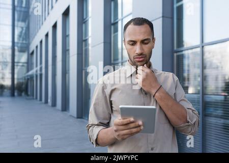 Seriöse und nachdenkliche afroamerikanische Geschäftsmann mit Tablet-Computer außerhalb des modernen Bürogebäudes, Mann liest Nachrichten. Stockfoto