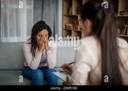 Traurige, weinende, schwarze, tausendjährige Patientin, die mit einem europäischen Arzt spricht, braucht eine Therapie im Klinikinneren Stockfoto