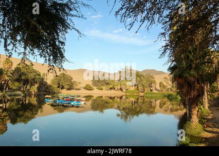 Huacachina - eine Oase mitten in der peruanischen Wüste, Blick auf Palmen und eine Lagune mit Booten und Sanddünen im Hintergrund. Stockfoto