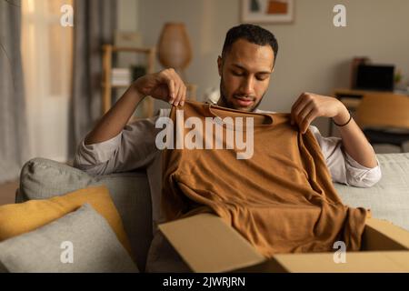 Hübscher junger schwarzer Kerl, der Pappkarton entpackt, versucht, einen gelieferten Kauf zu kaufen und neue Kleidung zu Hause herausnimmt Stockfoto