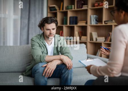 Verzweifelter, depressiver junger Kaukasier leidet an psychischen Problemen, hört sich eine schwarze Psychologin an Stockfoto