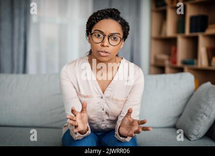 Seriöse junge schwarze Frau Arzt in Brille spricht und gestikuliert, Blick auf die Kamera im Büro Stockfoto