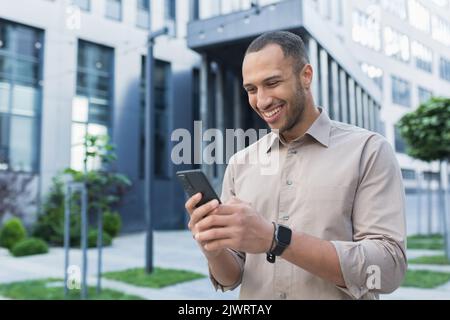 Junger afroamerikanischer Geschäftsmann mit Smartphone, lächelnd und glücklich beim Schreiben von Nachrichten online, Mann vor dem Bürogebäude beim Schreiben von Nachrichten an Freunde. Stockfoto