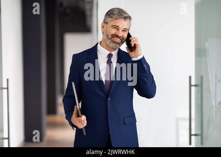 Lächelnder Geschäftsmann Im Mittleren Alter, Der Mit Dem Handy Spricht, Während Er Aus Dem Büro Geht Stockfoto