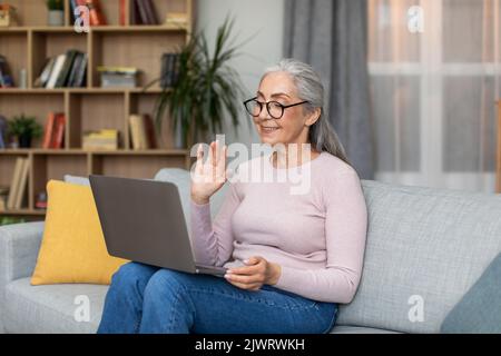 Lächelnde kaukasische ältere grauhaarige Frau mit Brille, die ihre Hand gegen die Laptop-Webcam im Wohnzimmer winkt Stockfoto