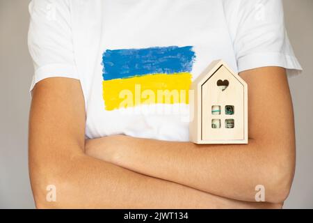 Ein Mädchen hält in den Händen ein kleines hölzernes Dekorationshaus mit Dollars, gekleidet in ein T-Shirt mit der Flagge der Ukraine, einem Flüchtling und einem Haus Stockfoto