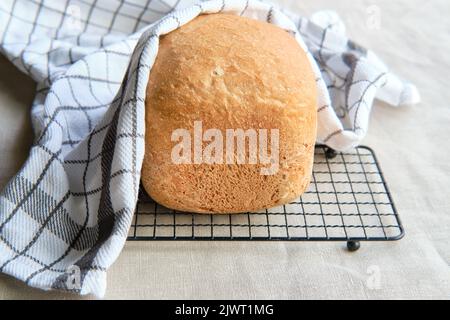 Hausgemachtes Bauernhaus gemischtes Brot. Nahaufnahme von Brot, das in der Backmaschine gebacken wurde. Vollkornbrot auf Trockengestell mit gestreifem Handtuch auf Textil Stockfoto