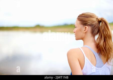 Gehe allein mit deinen Gedanken. Rückansicht einer jungen Frau, die beim Yoga auf einen See schaut. Stockfoto