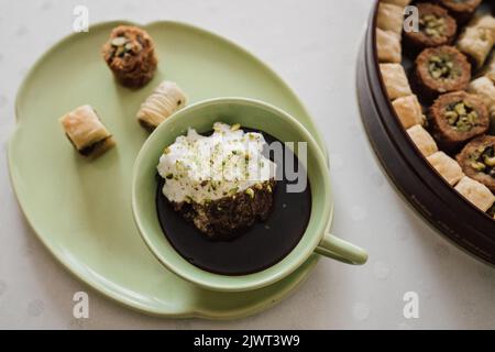 Trinkschokolade in passendem grünen Becher und Teller mit verschiedenen Baklava-Desserts auf weißer Tischdecke mit Polka-Punkt Stockfoto