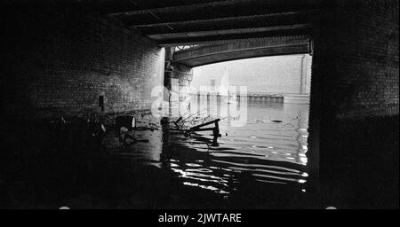 London, England, um 1967. Ein Blick unter einer Brücke über den Regent’s Canal. Zwei Jungen, die Mitglieder des Piratenclubs sind, segeln in einem kleinen Schlauchboot in der Ferne. Unter der Brücke hat sich eine große Menge an Schutt angesammelt. Der Pirate Club, ein Kinderbootclub, wurde 1966 in Gilbey's Wharf am Regents Canal in der Nähe von Camden, London, gegründet. Ihr Klubhaus war ein alter Lastkahn und eine Reihe kleiner Boote und Kanus wurden für die Kinder gespendet. Stockfoto