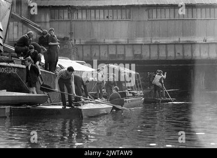 London, England, um 1967. Zwei Erwachsene Freiwillige und eine Gruppe Kinder des Piratenclubs an Bord von ‘Rosedale’, einem ausgedienten Lastkahn, der als Clubhaus diente. Andere Jungen Mann verschiedene Boote, Flöße und Kanus. Hinter ihnen befindet sich die Eisenbahnbrücke, die an die Gloucester Avenue angrenzt. Der Pirate Club, ein Kinderbootclub, wurde 1966 in Gilbey’s Wharf am Regent’s Canal in der Nähe von Camden, London, gegründet. Für die Kinder wurden eine Reihe kleiner Boote und Kanus gespendet. Stockfoto