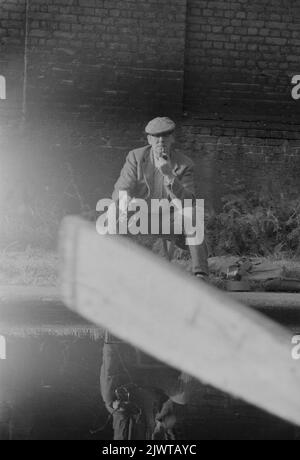 London, England, um 1967. Ein Angler, der eine Pfeife raucht, fischt vom Schlepptau auf dem Regent’s Canal in Camden zwischen Oval Road und Gloucester Road. Das Foto wurde von einem Boot des Piratenclubs aufgenommen. Im Vordergrund ist ein Ruder zu sehen. Der Pirate Club, ein Kinderbootclub, wurde 1966 in Gilbey’s Wharf am Regent’s Canal in der Nähe von Camden, London, gegründet. Ihr Klubhaus war ein alter Lastkahn und eine Reihe kleiner Boote und Kanus wurden für die Kinder gespendet. Stockfoto