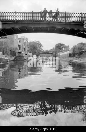 London, England, um 1967. Eine Familie, die auf der breiten Fußgängerbrücke über den Regent’s Canal im Cumberland Basin steht, beobachtet die Jungen des Pirates’ Club, die unter ihnen ein Boot paddeln. Im Regent’s Park befindet sich die Snowdon Aviary des Londoner Zoos. Der Pirates’ Club, ein Kinderbootclub, wurde 1966 in Gilbey’s Wharf am Regent’s Canal in der Nähe von Camden, London, gegründet. Ihr Klubhaus war ein alter Lastkahn und eine Reihe kleiner Boote und Kanus wurden für die Kinder gespendet. Stockfoto