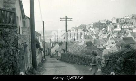 Cornwall 1950s. Ein malerischer Blick auf den Church Hill, Port Issac, ein kleines Fischerdorf an der Atlantikküste von Nord-Cornwall. Ein junges Mädchen steht im Vordergrund und blickt den steilen Hügel hinunter. Verschiedene Hütten und Gebäude, darunter das alte Schulhaus, liegen auf dem Hügel. Der Ozean ist in der Ferne sichtbar. Stockfoto