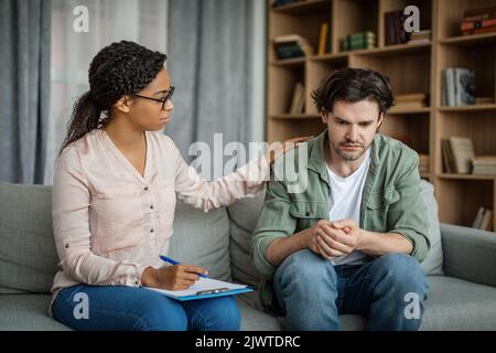 Serious junge schwarze Ärztin beruhigt europäischen Kerl, Client leidet an Depressionen im Büro Klinik Innenraum Stockfoto